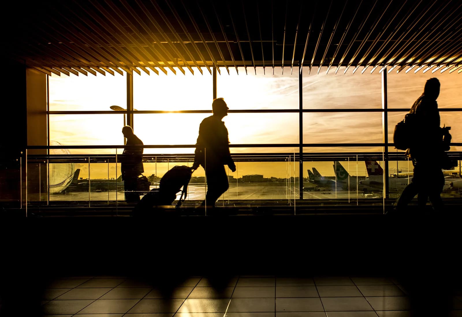 Travelers pulling suitcases through an airport terminal with aircraft outside at sunset.